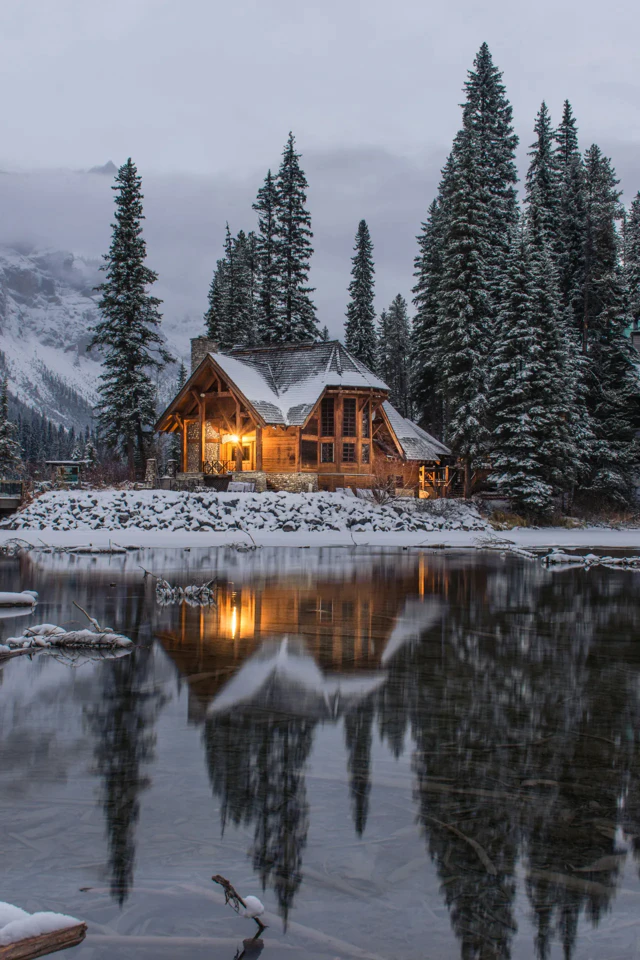 Chalet en bois éclairé au bord d'un lac entouré de sapins enneigés.