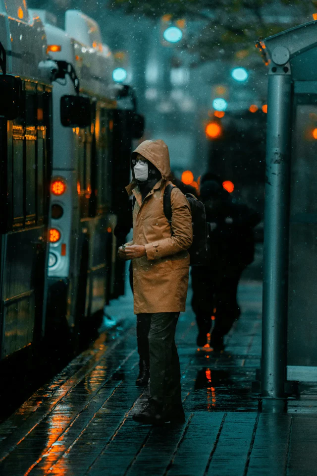 Personne en manteau beige et masque attendant un bus sous la pluie.