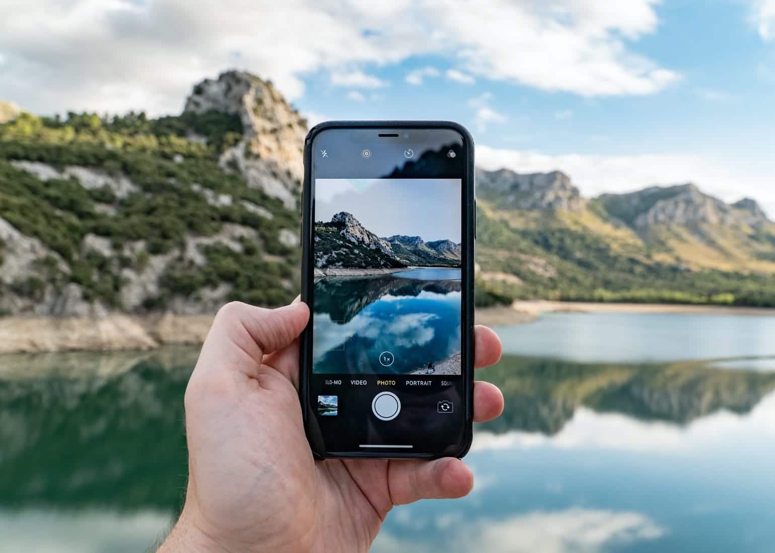 Main tenant un smartphone capturant une image de paysage montagneux avec lac en arrière-plan.