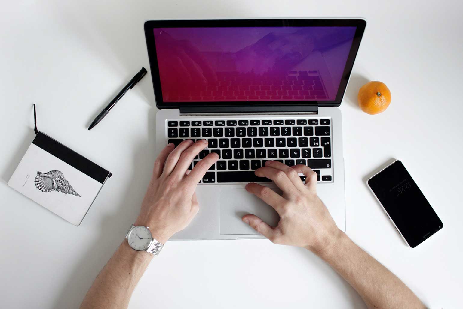 Mains tapant sur un ordinateur portable avec un carnet, un stylo, une orange et un smartphone sur un bureau blanc