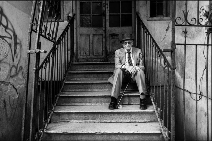 Un homme âgé en costume et chapeau assis sur des marches devant une porte ancienne.