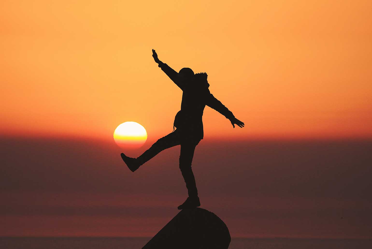 Personne en équilibre sur un rocher au coucher du soleil avec des bras étendus.