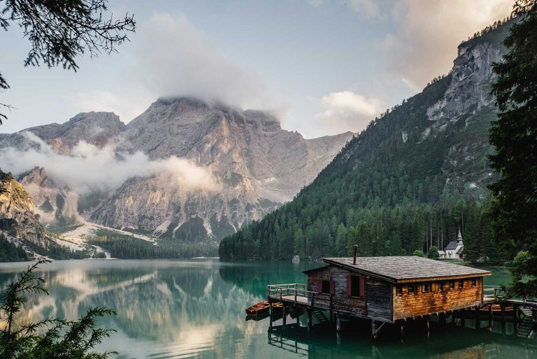 Cabane en bois au bord d'un lac entouré de montagnes et forêts avec des nuages bas.
