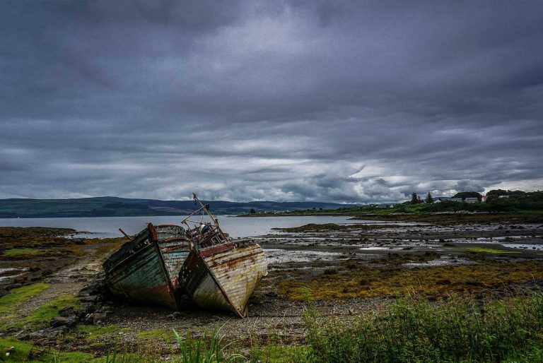 Deux bateaux en bois abandonnés sur une côte rocheuse sous un ciel nuageux.
