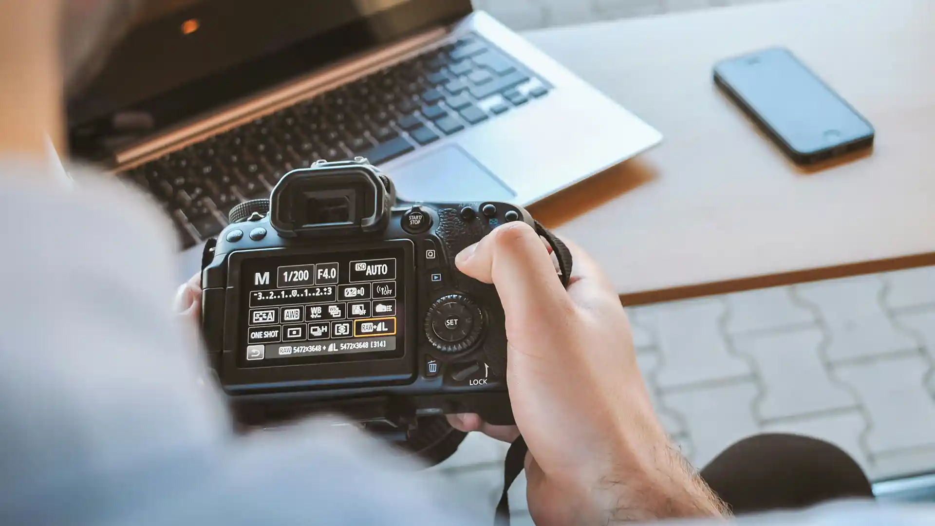 Personne tenant un appareil photo reflex numérique devant un ordinateur portable et un smartphone sur une table.