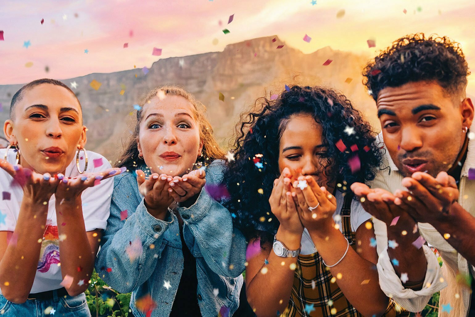 Groupe de personnes soufflant des confettis colorés sous un ciel de coucher de soleil.