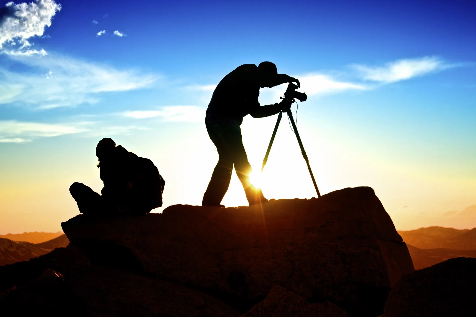 Deux photographes en silhouette au sommet d'une montagne au coucher du soleil, l'un derrière son trépied