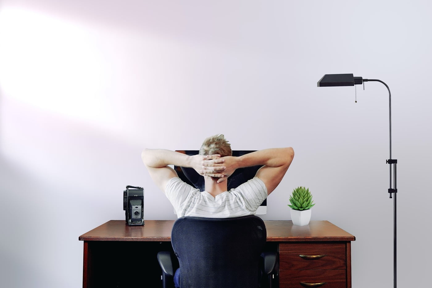 Personne assise à un bureau, vue de dos, devant un ordinateur avec les mains croisées derrière la tête.
