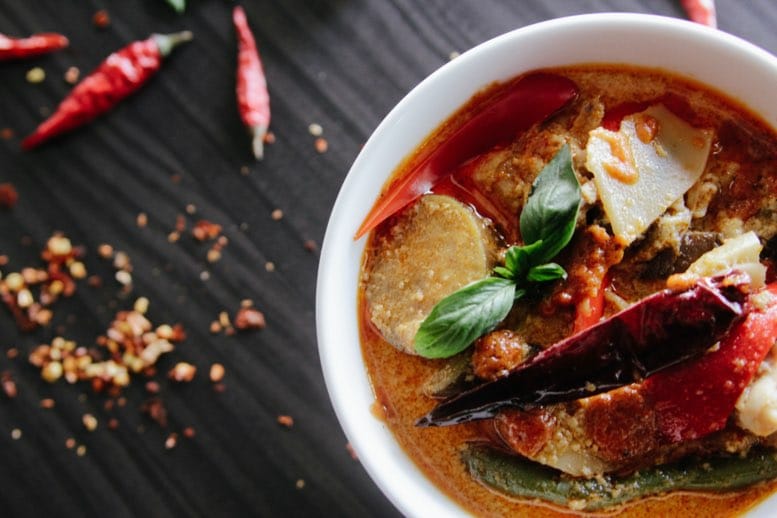 Bol de curry épicé avec des légumes décoré de piments rouges et de feuilles de basilic sur une table foncée.