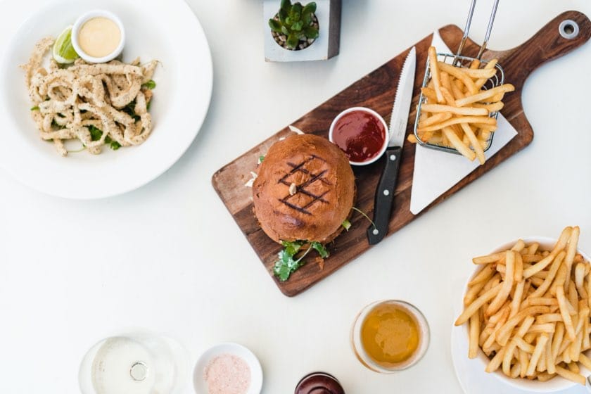Burger gourmet avec frites servies sur une planche en bois entourée d'autres plats et boissons sur une table blanche.