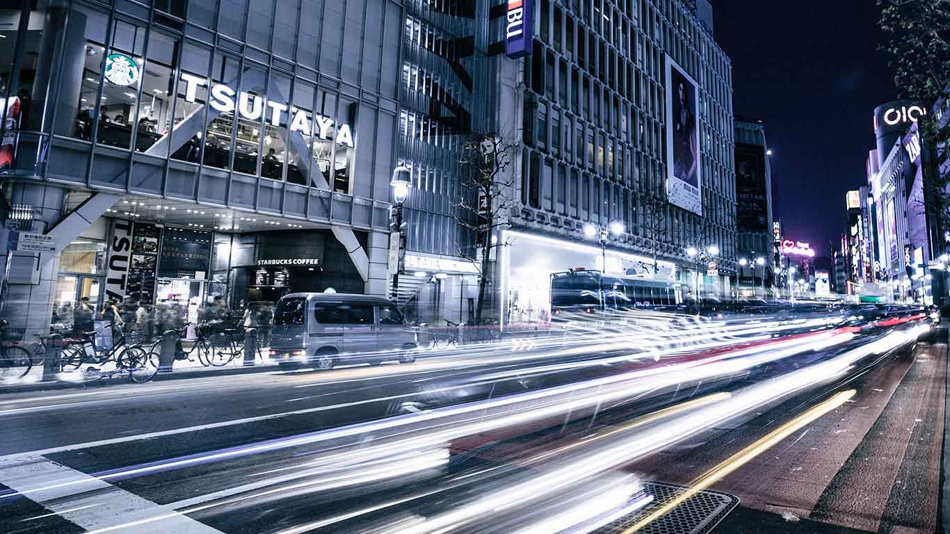 Scène nocturne urbaine animée avec des traînées lumineuses de phares de voiture et des bâtiments éclairés